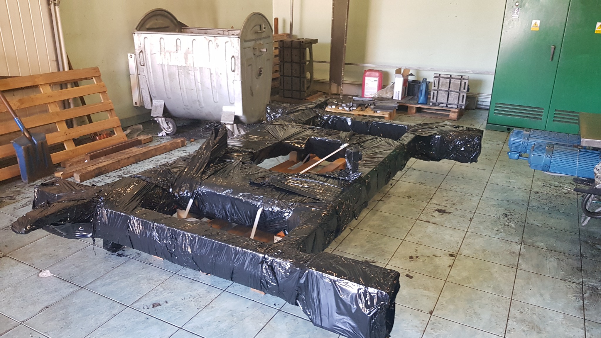 Metalowa konstrukcja, prawdopodobnie rama urządzenia, owinięta czarną folią ochronną, leżąca na podłodze warsztatu. W tle widoczny kontener na odpady, palety i maszyny.