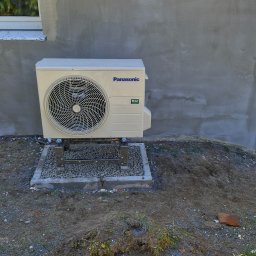 Tech Power Systemy Instalacyjne Daniel Łakomiak - Beżowa jednostka zewnętrzna klimatyzacji Panasonic na metalowym stelażu, zamontowana przy szarej ścianie budynku. Widoczne okno i gruz na podwórku.