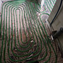 Tech Power Systemy Instalacyjne Daniel Łakomiak - Zielone rury ogrzewania podłogowego ułożone na folii izolacyjnej w surowym pomieszczeniu z betonowymi ścianami. Widoczny fragment instalacji przy ścianie.