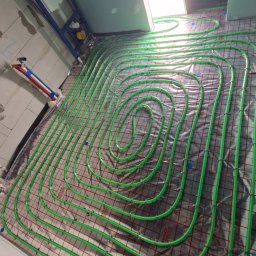 Tech Power Systemy Instalacyjne Daniel Łakomiak - Układanie zielonych rur ogrzewania podłogowego na folii z czerwoną siatką. Widoczny fragment instalacji WC podtynkowej i fragment osoby w dolnej części kadru.