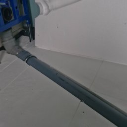 Tech Power Systemy Instalacyjne Daniel Łakomiak - Szara rura kanalizacyjna podłączona do niebieskiej ramy stelaża WC, widoczna na tle białej ściany i podłogi z płyt styropianowych.