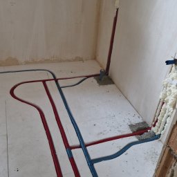 Tech Power Systemy Instalacyjne Daniel Łakomiak - Instalacja ogrzewania podłogowego w trakcie montażu: czerwone i niebieskie rury ułożone na białej izolacji, pianka montażowa przy ścianie. Widok z góry.