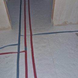 Tech Power Systemy Instalacyjne Daniel Łakomiak - Instalacja wodna i kanalizacyjna w surowym stanie: czerwone i niebieskie rury prowadzone w podłodze, stelaż podtynkowy WC z szarą rurą kanalizacyjną.