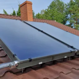 Dwa kolektory słoneczne zamontowane na brązowym dachu z dach&oacute;wki ceramicznej, z widocznym kominem z cegły i zielenią w tle. Instalacja z izolowanymi przewodami.