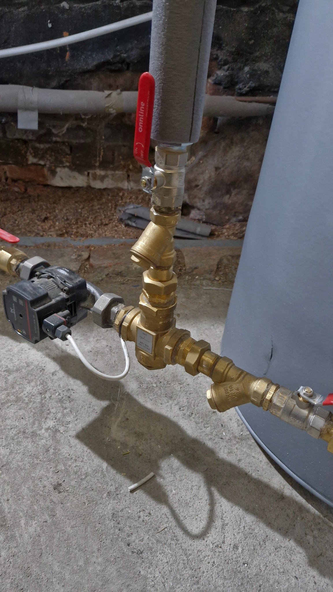 Złożona instalacja hydrauliczna z mosiężnymi kształtkami i pompą obiegową, widoczna na tle surowej ściany i betonowej podłogi. Czerwone zawory odcinające.