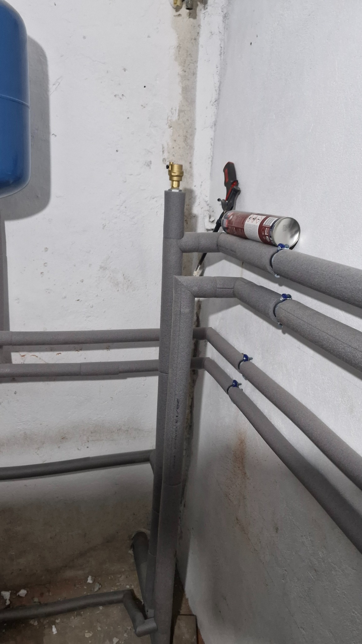 Szare, zaizolowane rury grzewcze zamontowane wzdłuż ściany. Widoczny zbiornik wyrównawczy i puszka z pianką montażową. Instalacja w surowym pomieszczeniu.