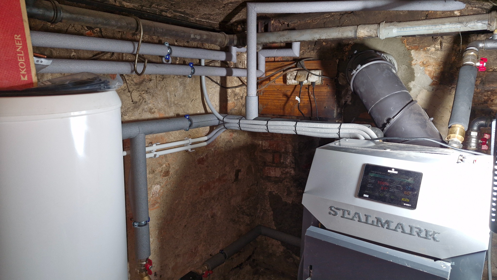 Złożona instalacja grzewcza z kotłem Stalmark, licznymi rurami w izolacji i zbiornikiem. Widoczne elementy montażu i podłączenia w piwnicy.