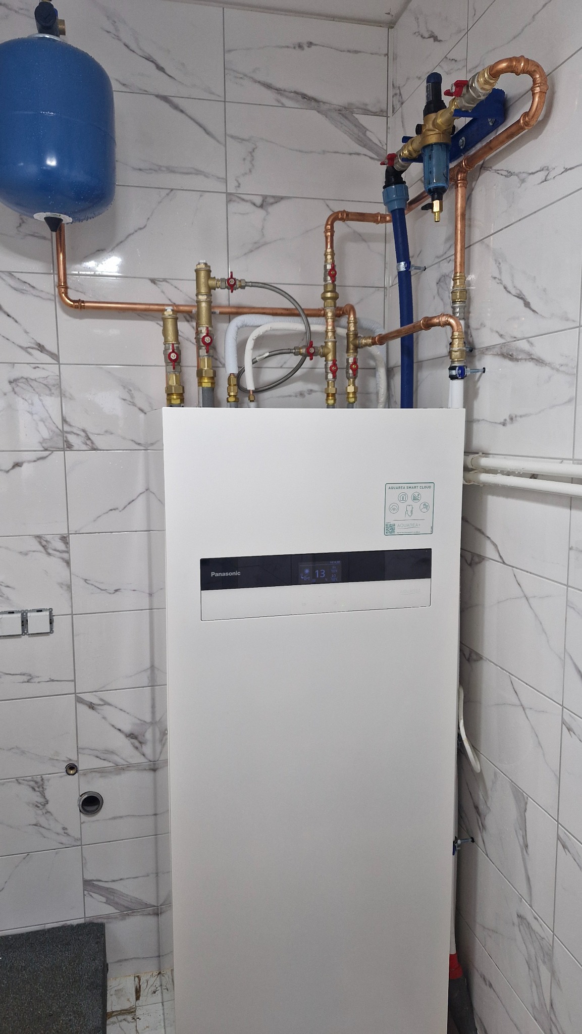 Zmontowana, biała pompa ciepła Panasonic z widocznymi miedzianymi rurami i zaworami na tle marmurowych płytek. Widoczny zbiornik wyrównawczy.