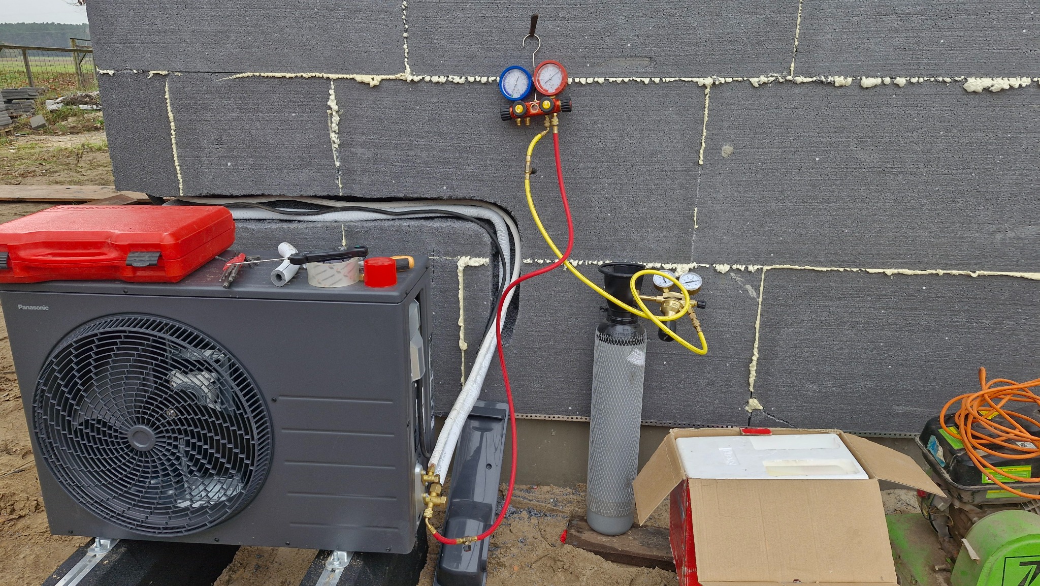 Szara pompa ciepła Panasonic, podłączona do manometru i butli z czynnikiem chłodniczym, ustawiona przy ścianie budynku z szarego bloczka. Widoczne narzędzia i akcesoria.