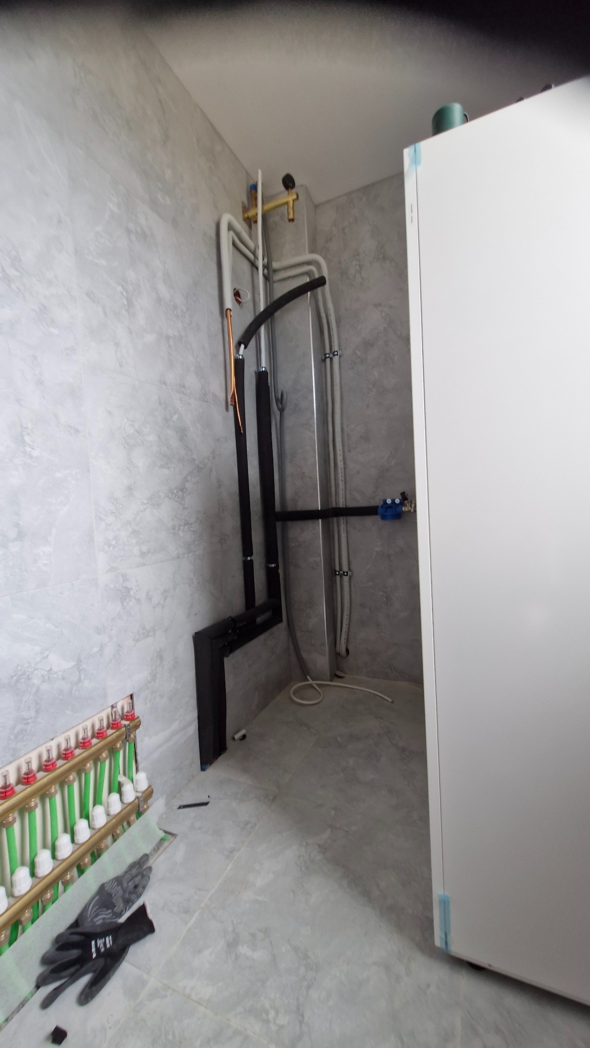 Szara łazienka w trakcie instalacji ogrzewania podłogowego: widoczny rozdzielacz, rury w izolacji i robocze rękawice na podłodze.