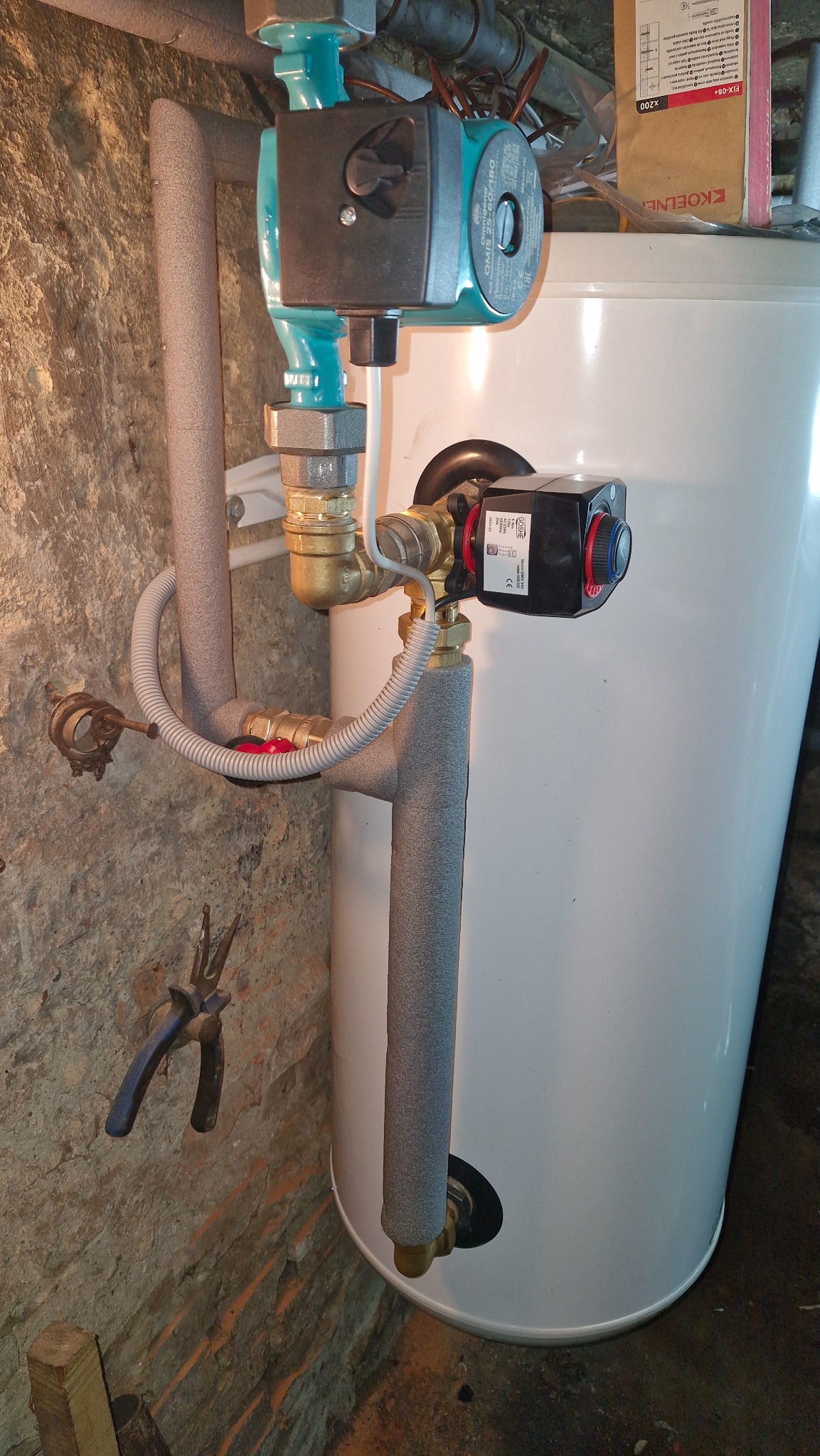Instalacja centralnego ogrzewania z pompą obiegową i zaworem termostatycznym, rury zaizolowane, szczypce na ścianie z cegły, zbiornik biały.