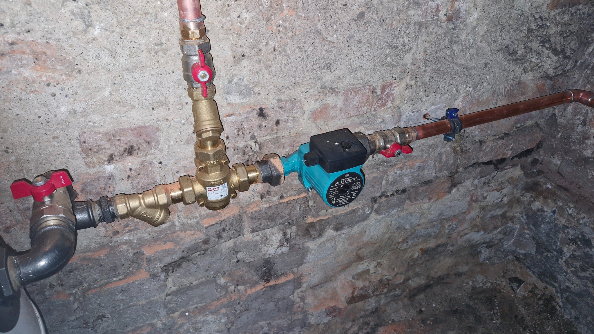 Fragment instalacji CO z pompą obiegową Omnigena Omis 25-60/180, zaworami kulowymi z czerwonymi rączkami i filtrem skośnym na tle ściany z cegły.