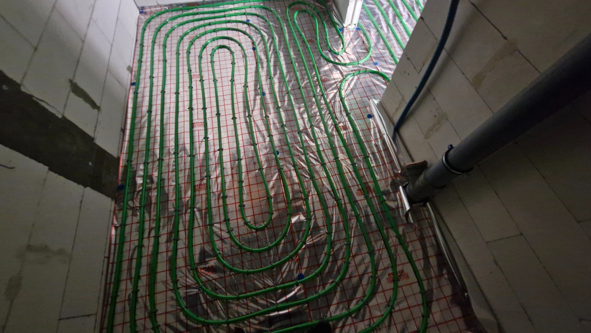 Zielone rury ogrzewania podłogowego ułożone na folii izolacyjnej w surowym pomieszczeniu z betonowymi ścianami. Widoczny fragment instalacji przy ścianie.