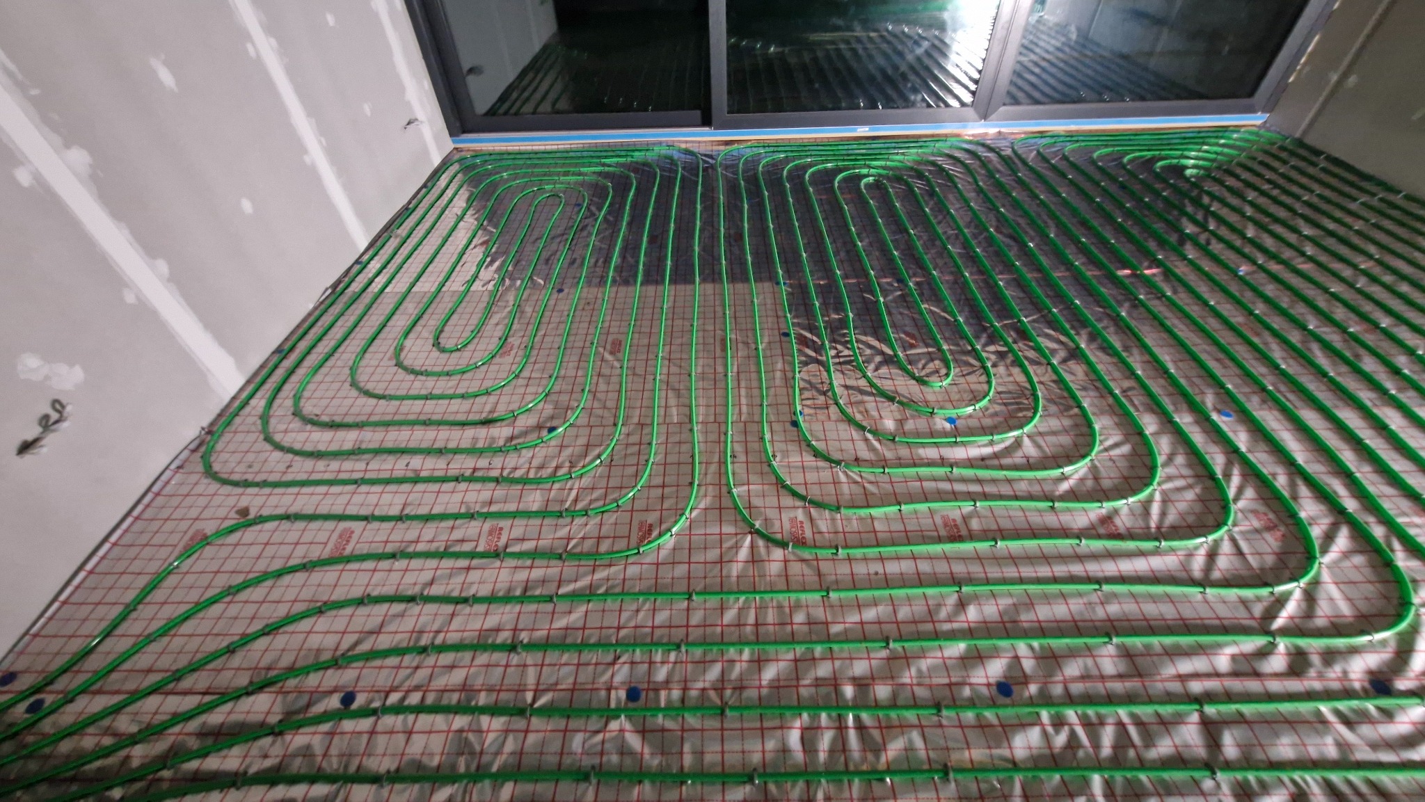 Zielone rury ogrzewania podłogowego ułożone na folii z siatką, widok z góry, przygotowane do zalania posadzką w nowym budynku z oknem dachowym.