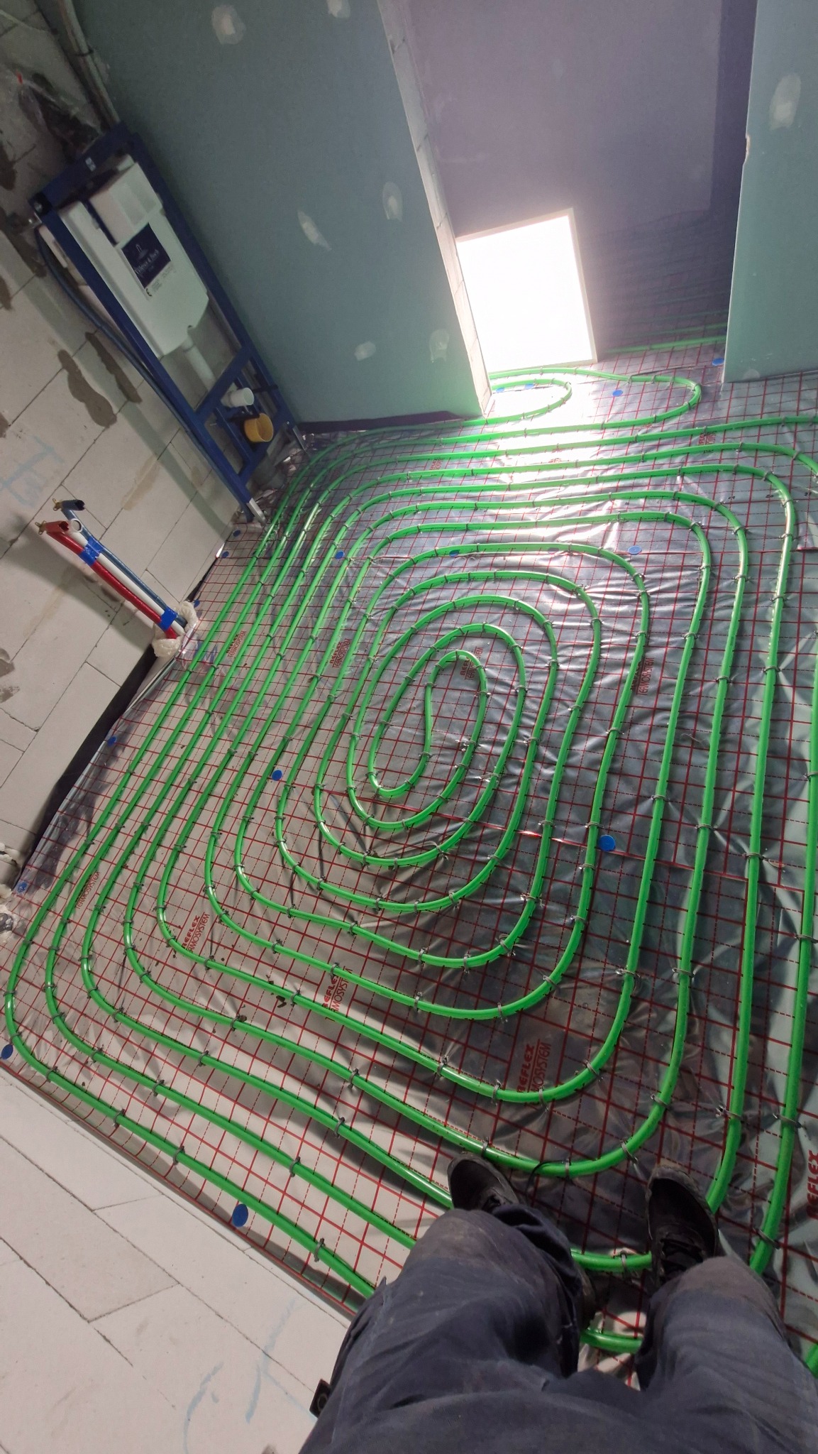 Układanie zielonych rur ogrzewania podłogowego na folii z czerwoną siatką. Widoczny fragment instalacji WC podtynkowej i fragment osoby w dolnej części kadru.