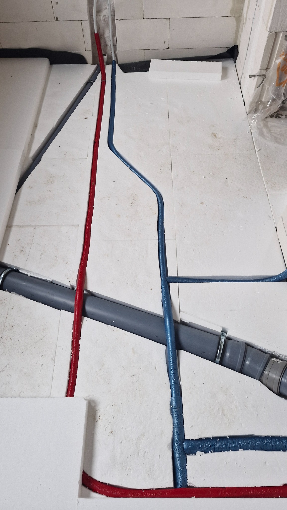 Układanie instalacji ogrzewania podłogowego: czerwone i niebieskie rury w izolacji na białym styropianie, szara rura kanalizacyjna. Widok z góry, etap prac.