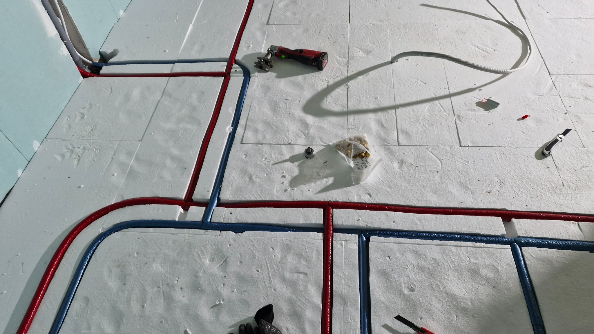 Rozłożone rury ogrzewania podłogowego (czerwone i niebieskie) na białym styropianie. Widoczne narzędzia: obcinak, nożyk, złączki. Kąt z góry.