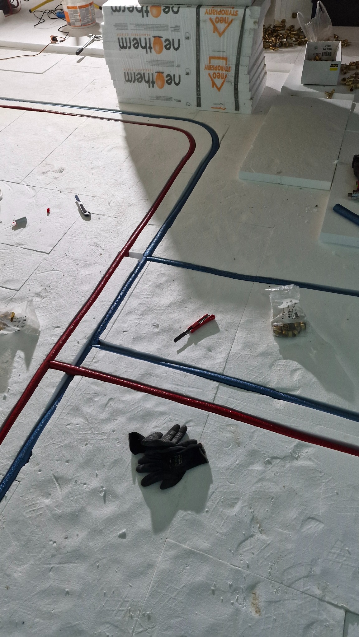 Instalacja ogrzewania podłogowego: czerwone i niebieskie rury ułożone na białym styropianie, czarne rękawice robocze, nożyk i akcesoria instalacyjne w tle.