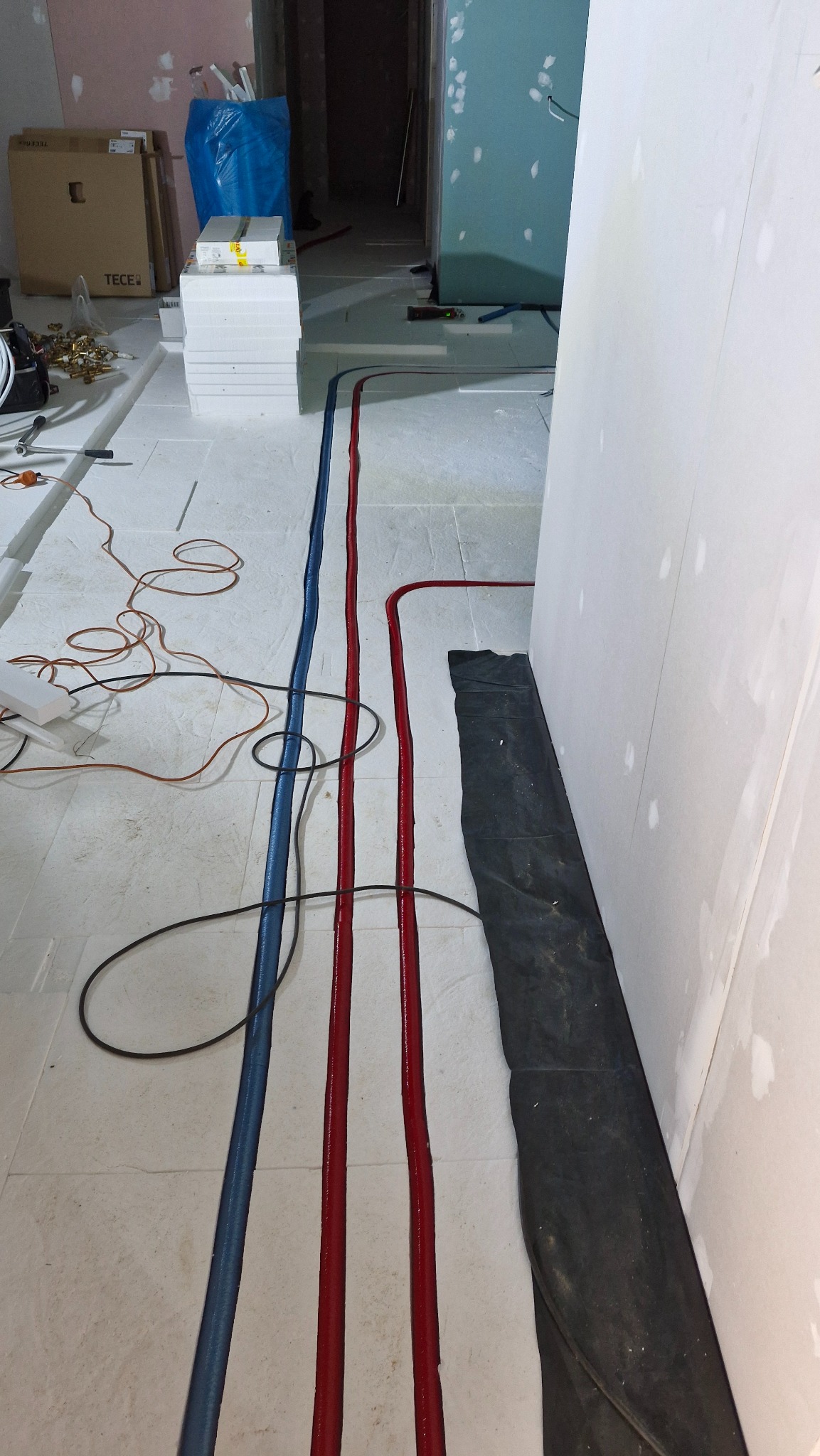 Instalacja ogrzewania podłogowego w trakcie montażu: rury grzewcze (czerwone i niebieskie) ułożone na izolacji, przygotowane do zalania posadzką. Widoczne materiały instalacyjne.