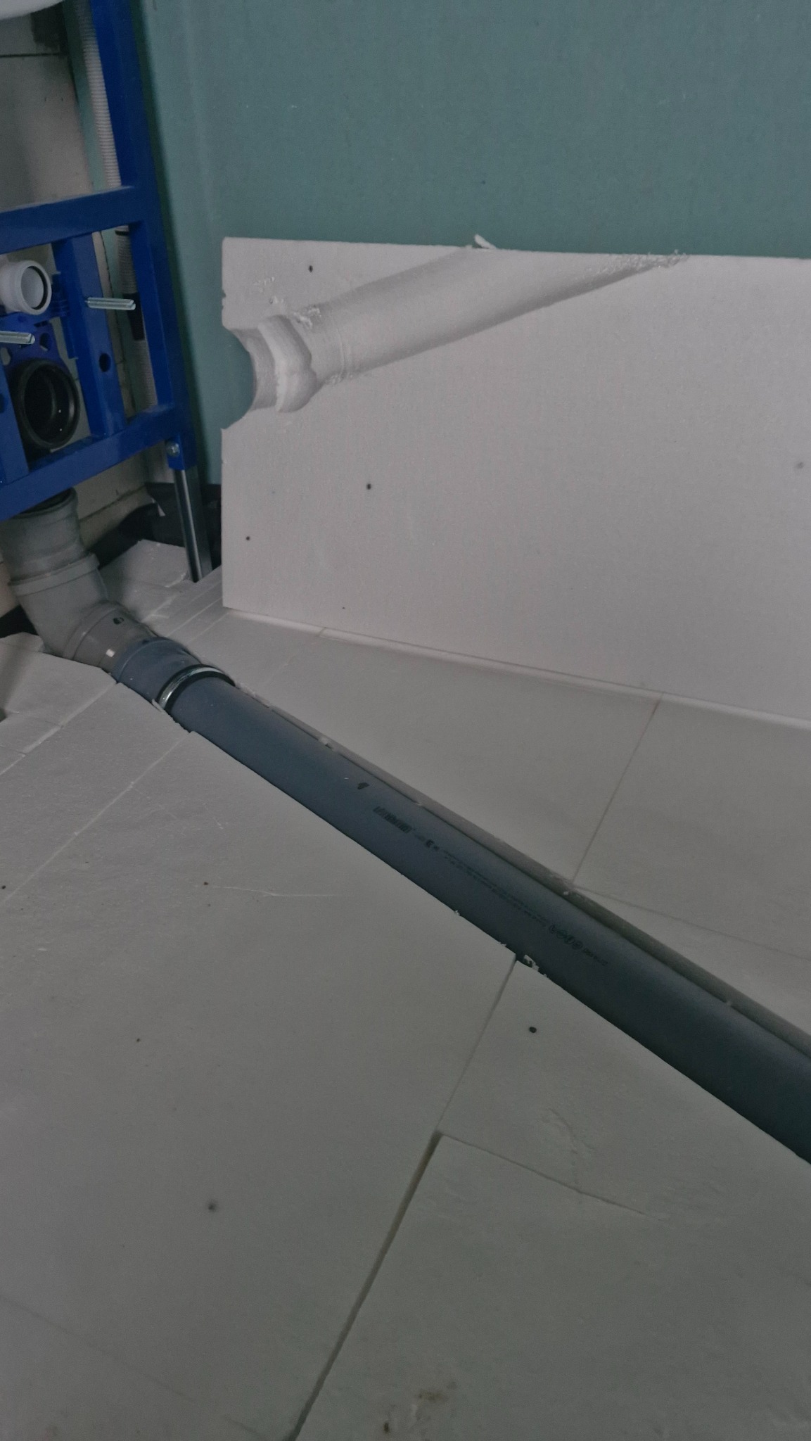 Szara rura kanalizacyjna podłączona do niebieskiej ramy stelaża WC, widoczna na tle białej ściany i podłogi z płyt styropianowych.