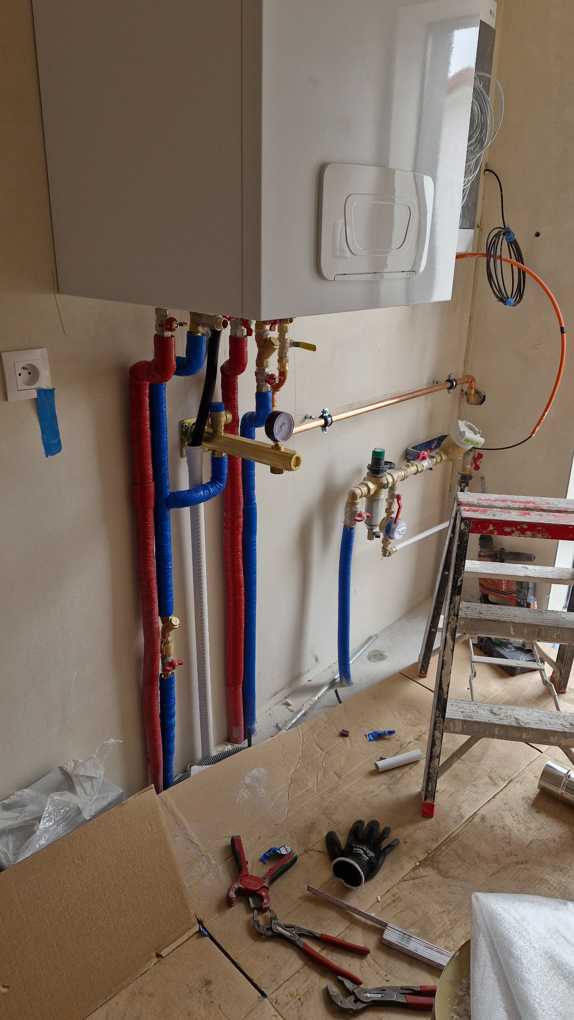 Biały piec gazowy zamontowany na ścianie, z podłączonymi rurami w izolacji czerwonej i niebieskiej. Na podłodze narzędzia i drabina.