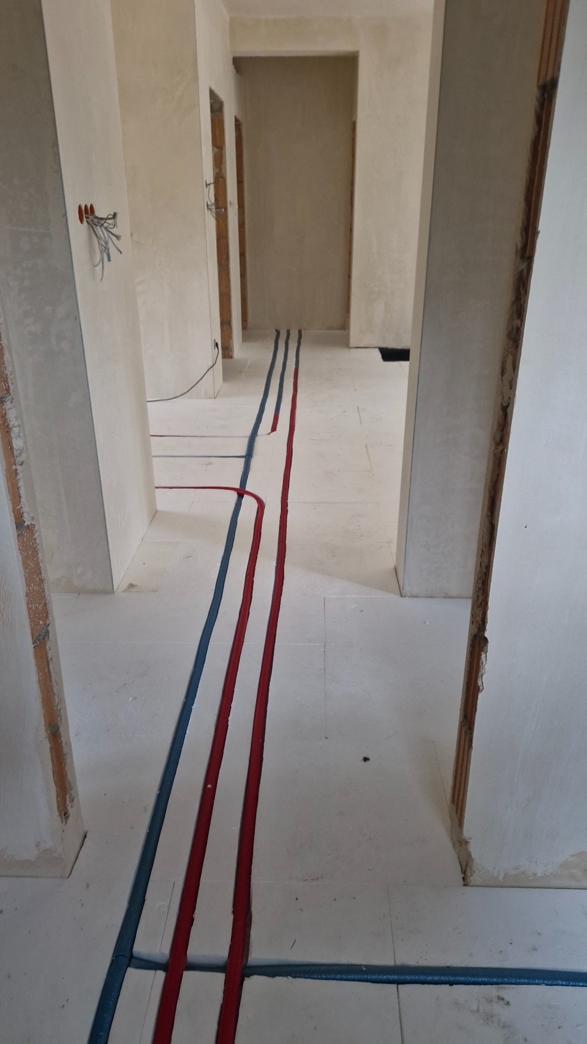 Instalacja ogrzewania podłogowego w trakcie montażu: czerwone i niebieskie rury prowadzone wzdłuż korytarza na białej powierzchni podłogi. Widoczne kable elektryczne.