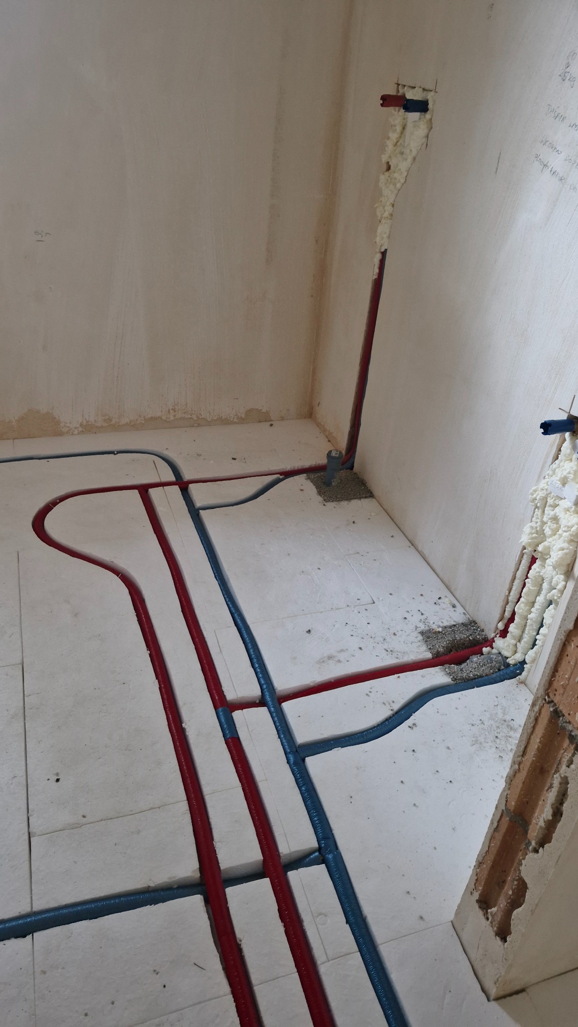 Instalacja ogrzewania podłogowego w trakcie montażu: czerwone i niebieskie rury ułożone na białej izolacji, pianka montażowa przy ścianie. Widok z góry.