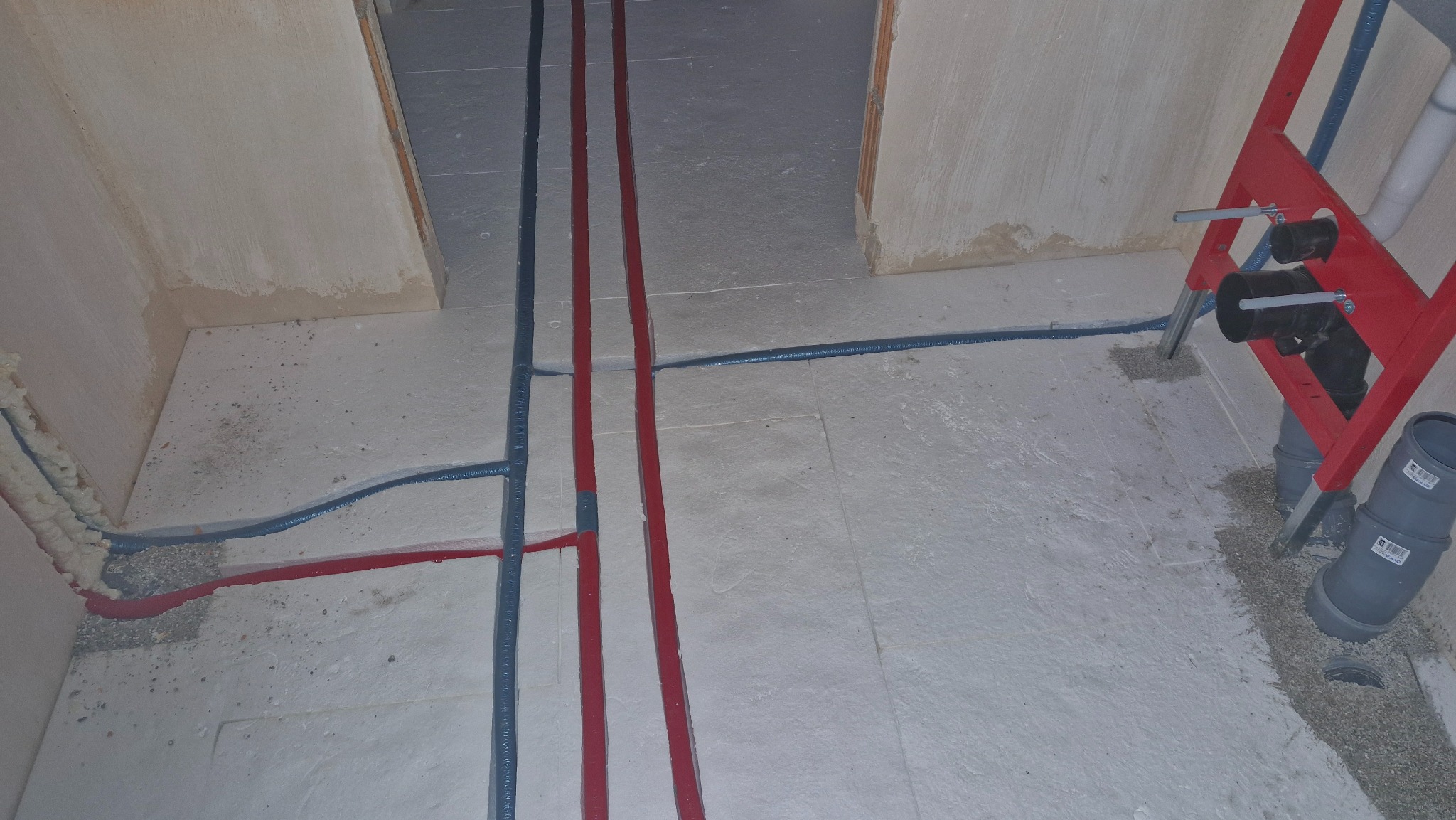 Instalacja wodna i kanalizacyjna w surowym stanie: czerwone i niebieskie rury prowadzone w podłodze, stelaż podtynkowy WC z szarą rurą kanalizacyjną.