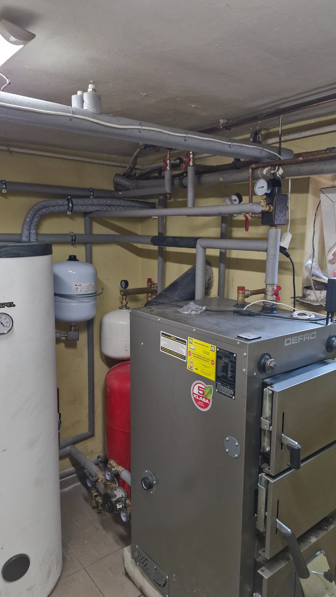 Kotłownia z kotłem Defro, zbiornikiem buforowym i instalacją CO z rurami w izolacji. Widoczne manometry i zawory. Całość w pomieszczeniu o żółtych ścianach.
