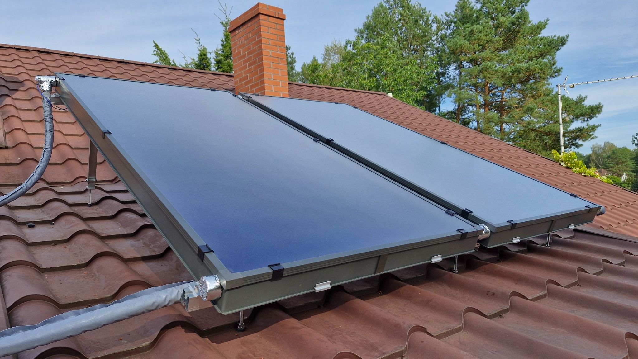 Dwa kolektory słoneczne zamontowane na brązowym dachu z dachówki ceramicznej, z widocznym kominem z cegły i zielenią w tle. Instalacja z izolowanymi przewodami.