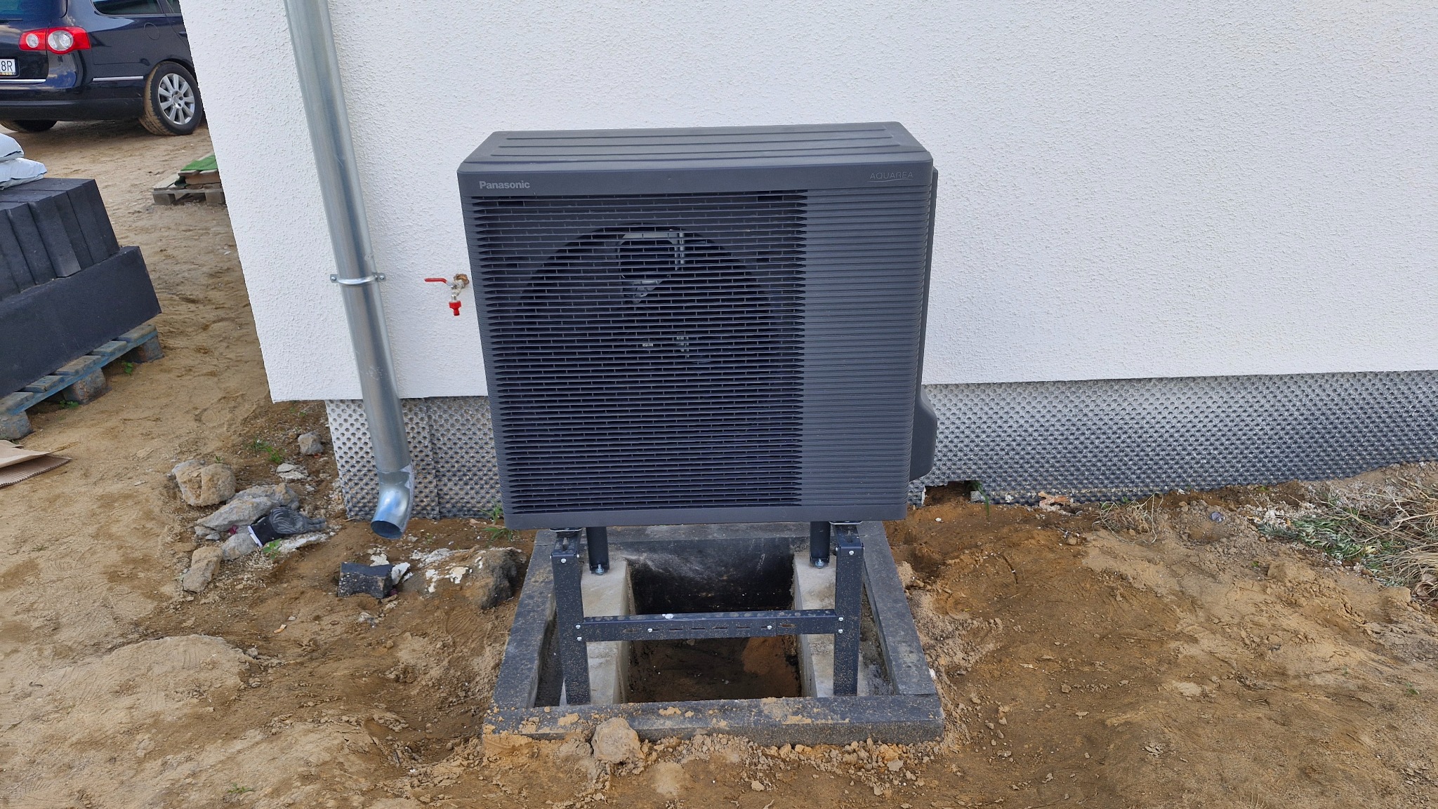 Szara pompa ciepła Panasonic Aquarea zamontowana na betonowej podstawie przy ścianie budynku z rurą spustową. Widoczny fragment samochodu i materiały budowlane.