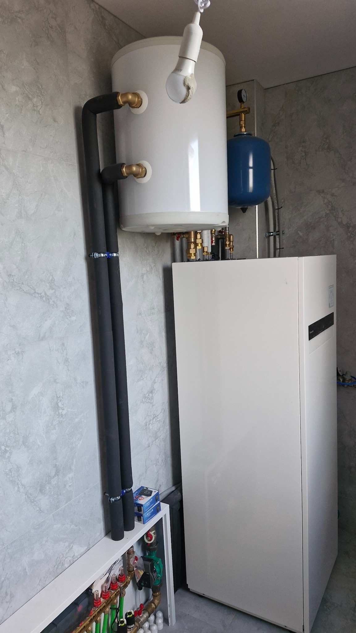 Biały bojler i pompa ciepła z czarnymi rurami w szarej łazience. Widoczne zawory i manometr. Elementy instalacji grzewczej na tle marmurowych płytek.