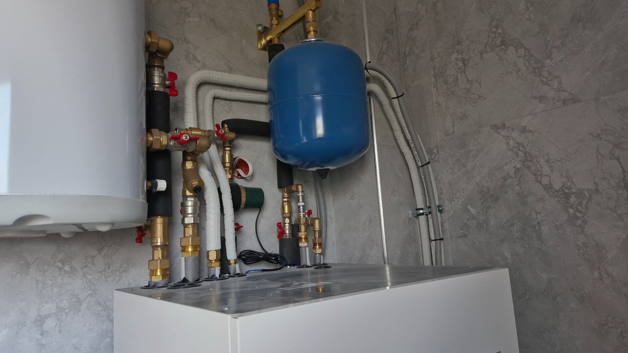Złożona instalacja grzewcza z białym zbiornikiem, niebieskim naczyniem wzbiorczym i licznymi rurami, zaworami, zamontowana na tle ściany z szarej marmurowej płytki.
