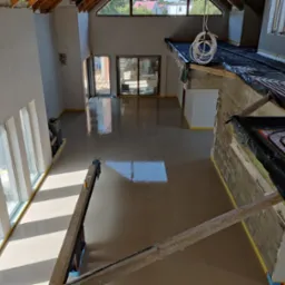 Świeżo wylana, mokra posadzka w surowym wnętrzu z oknami, widoczne ogrzewanie podłogowe w części pomieszczenia, zabezpieczone ściany taśmą malarską.