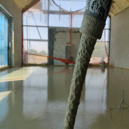 Anhi-Tech Patryk Drobnikowski - Wylewka samopoziomująca rozprowadzana w nowym budynku z dużymi oknami, widoczne narzędzie do rozprowadzania masy.