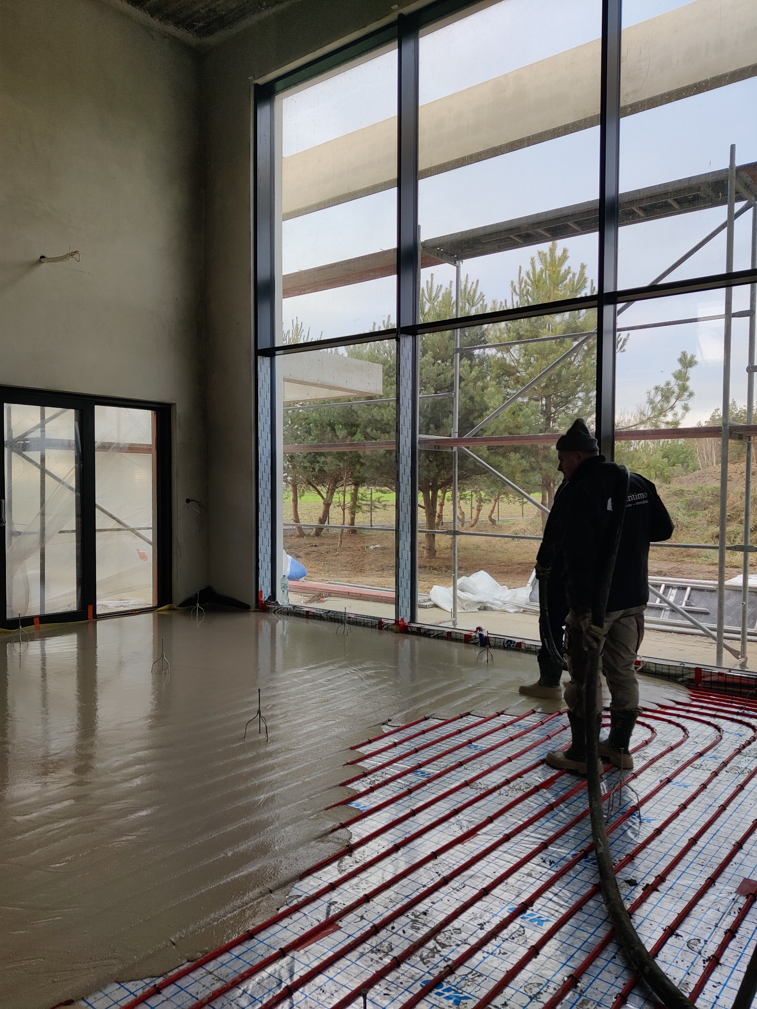 Ekipa w trakcie wylewania posadzki na ogrzewanie podłogowe w przestronnym wnętrzu z dużymi oknami, widoczne szare ściany w surowym stanie.