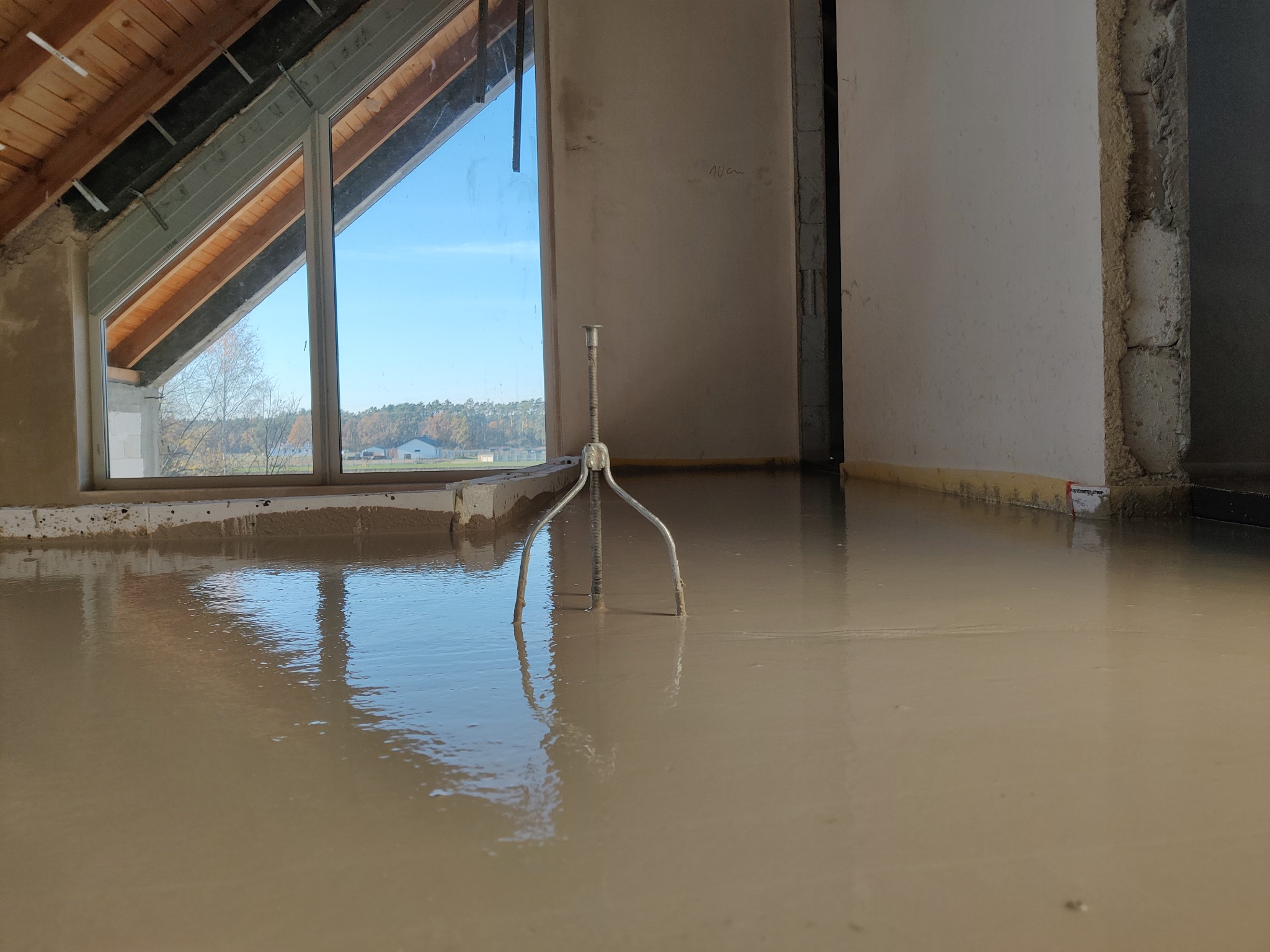 Świeżo wylana, mokra posadzka w pomieszczeniu z oknem dachowym, widoczny trójnóg do rozprowadzania wylewki.