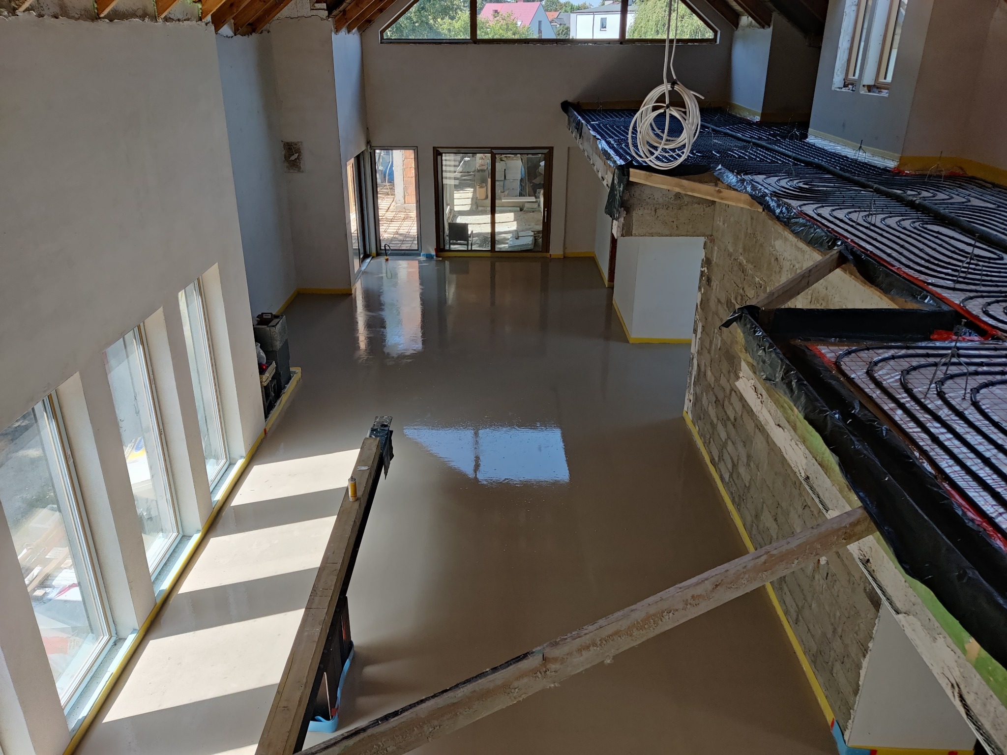 Świeżo wylana, mokra posadzka w surowym wnętrzu z oknami, widoczne ogrzewanie podłogowe w części pomieszczenia, zabezpieczone ściany taśmą malarską.
