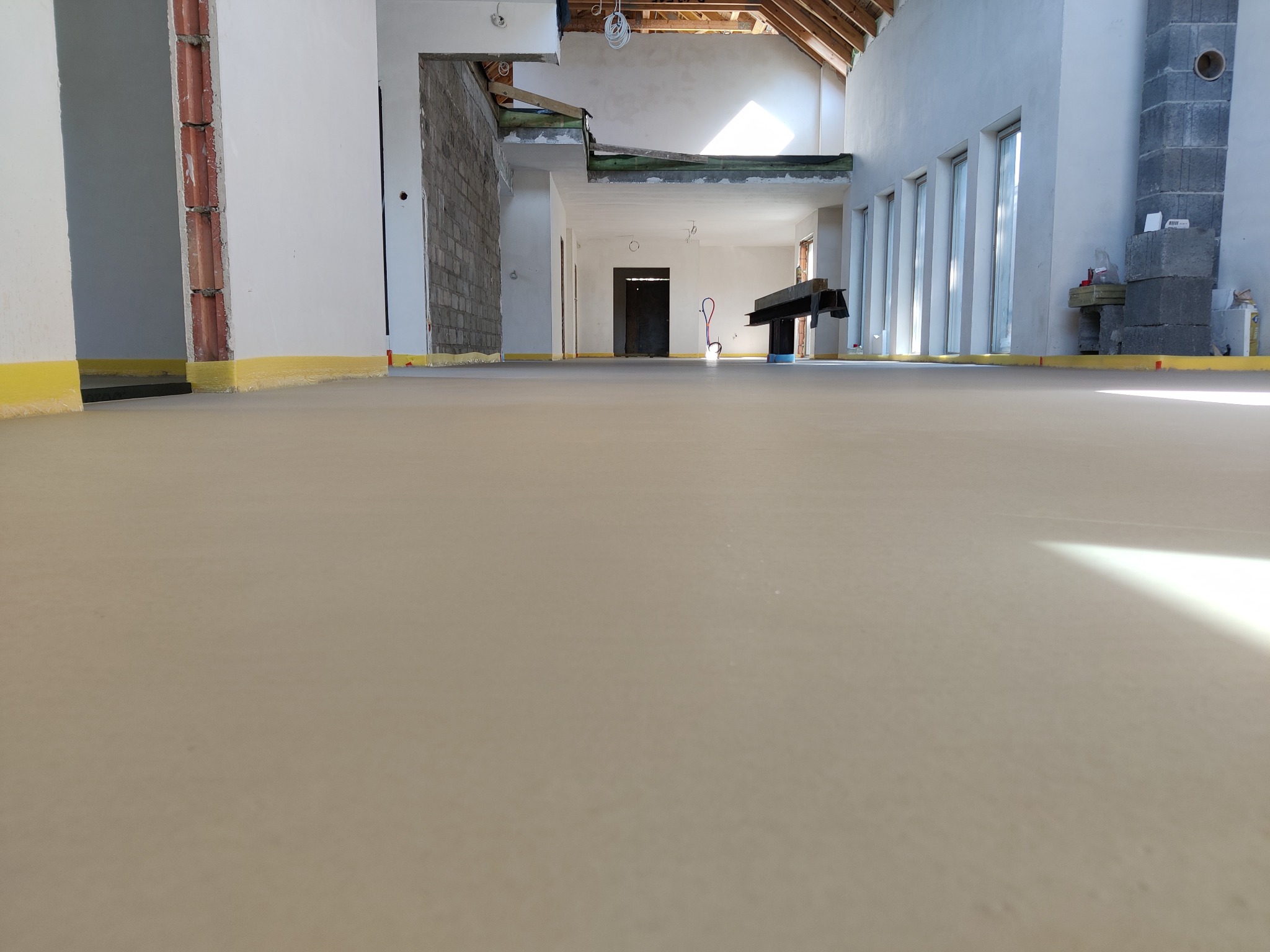 Świeżo wylana, jasna posadzka w przestronnym wnętrzu w trakcie budowy, widoczne żółte taśmy zabezpieczające przy ścianach, surowy komin z szarych pustaków i drewniana konstrukcja dachu.