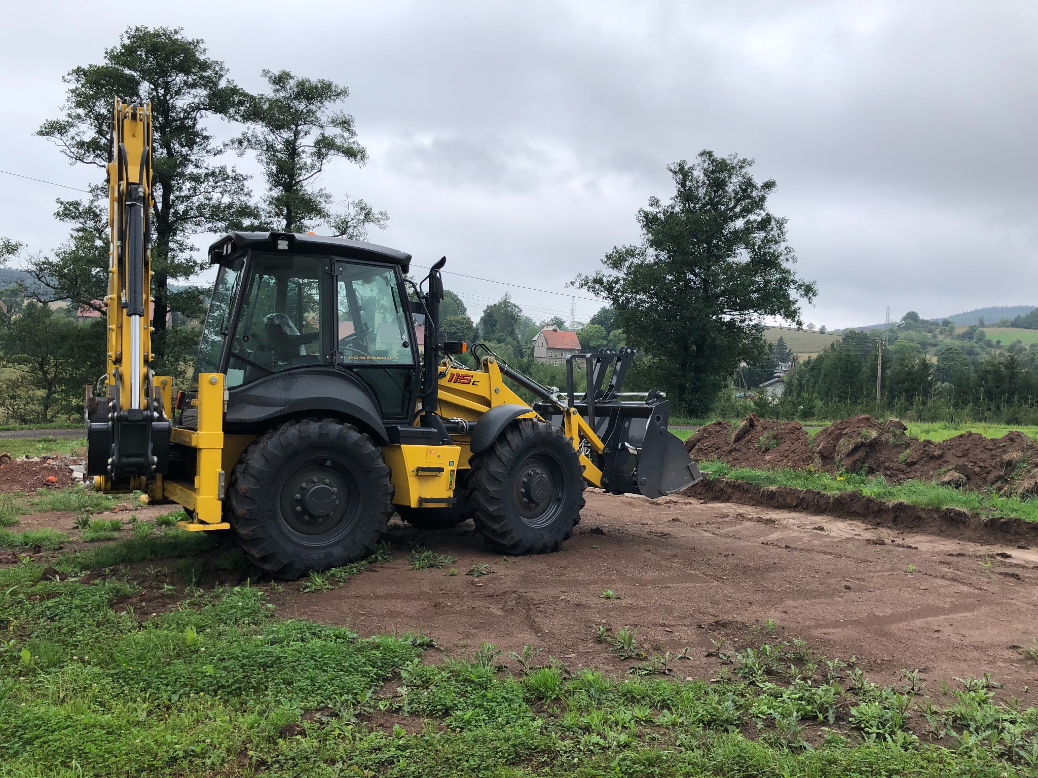 Żółto-czarna koparko-ładowarka 115C na placu budowy z wykopanym rowem w tle, widok z boku w pochmurny dzień.