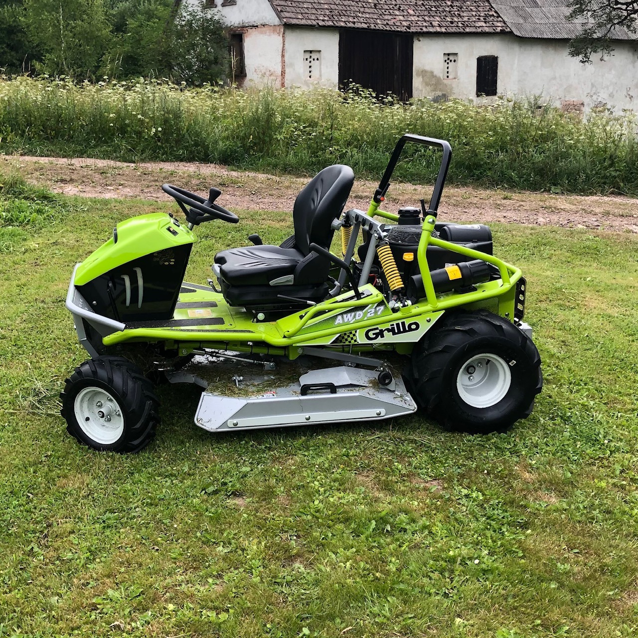 Zielona kosiarka samojezdna Grillo AWD 27 na trawniku przed białym budynkiem gospodarczym z ciemnymi drzwiami i oknami.