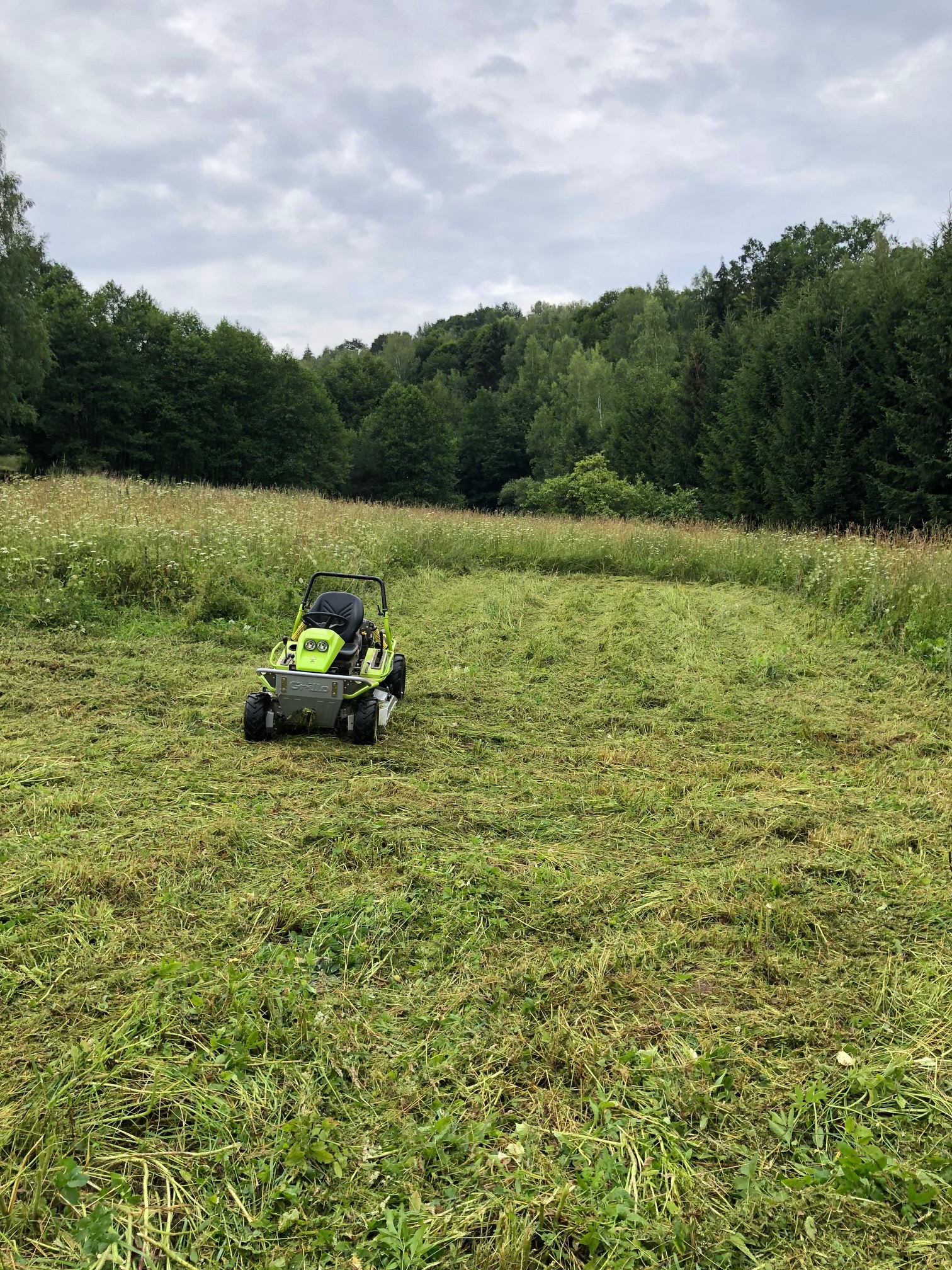 Zielona kosiarka samojezdna z operatorem na nierównym, częściowo skoszonym terenie z wysoką trawą i polnymi kwiatami, otoczonym lasem pod pochmurnym niebem.