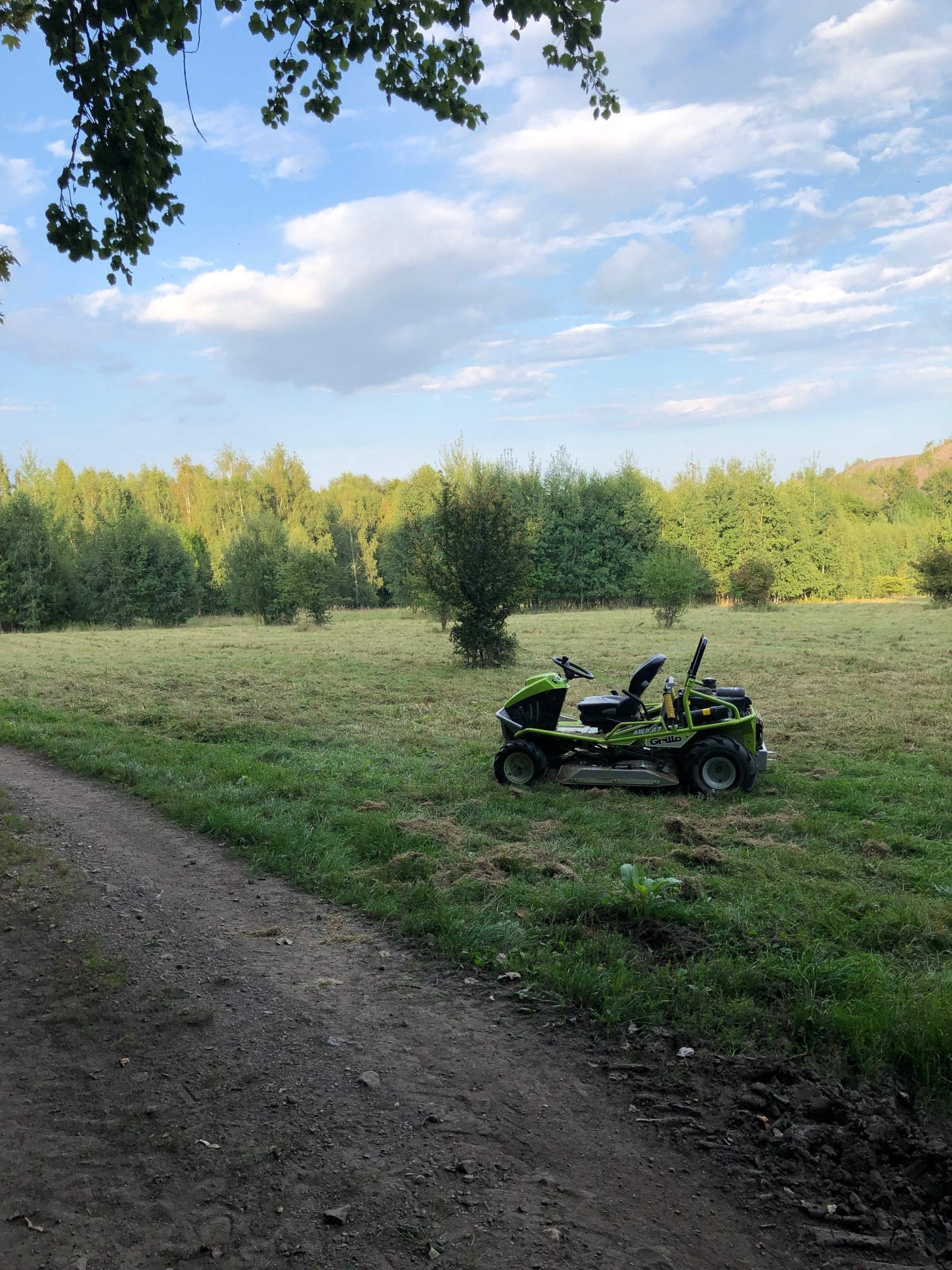 Zielona kosiarka samojezdna Grillo AWD 27 zaparkowana na skraju skoszonego trawnika, z widocznym fragmentem polnej drogi i drzewami w tle, w słoneczny dzień.
