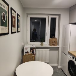 VS Wykończenie wnętrz Viktor Semenov - Wnętrze kuchni po remoncie: szare ściany, marmurowe płytki podłogowe, okrągły biały stół z wiklinowymi krzesłami, plakat na ścianie, torba na parapecie, widoczny nowy sprzęt AGD.