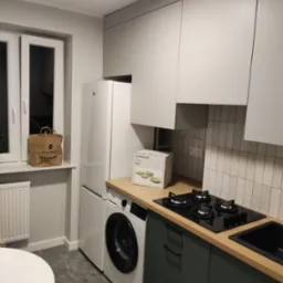 Nowoczesna kuchnia w odcieniach szarości i zieleni z drewnianym blatem, płytą grzewczą i zlewem, z pralką i lodówką w zabudowie, torba na oknie i pudełko z naczyniami na blacie.