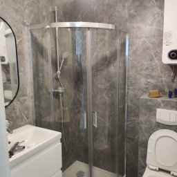 VS Wykończenie wnętrz Viktor Semenov - Nowoczesna łazienka z szarymi marmurkowymi płytkami, kabiną prysznicową z zaokrąglonymi drzwiami, białą umywalką z szafką i otwartą toaletą. Widoczne narzędzia na umywalce sugerują trwający montaż.