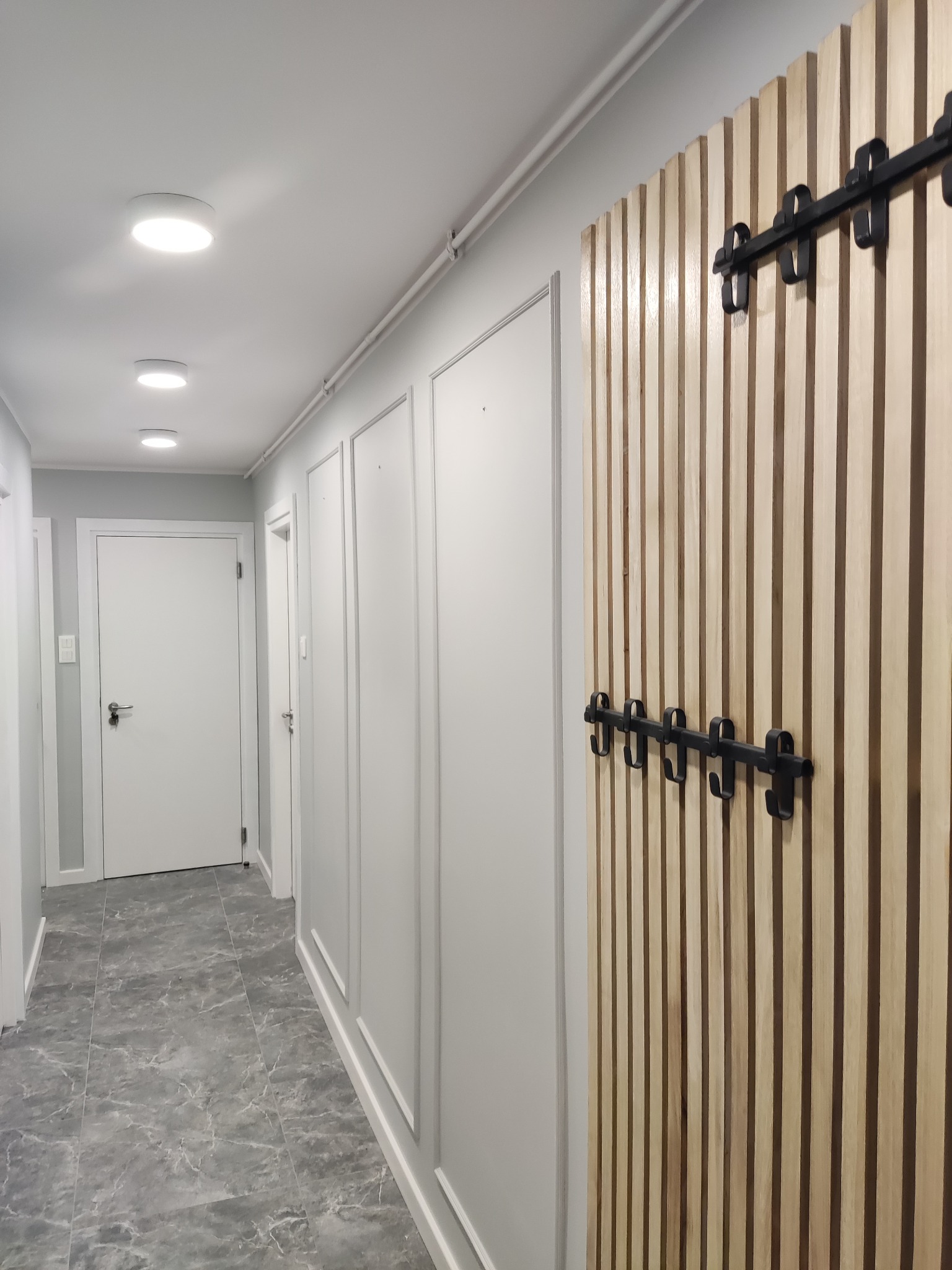 Nowoczesny przedpokój z szarą marmurową podłogą, białymi ścianami z dekoracyjnymi listwami i drewnianym panelem ściennym z czarnymi haczykami na ubrania.