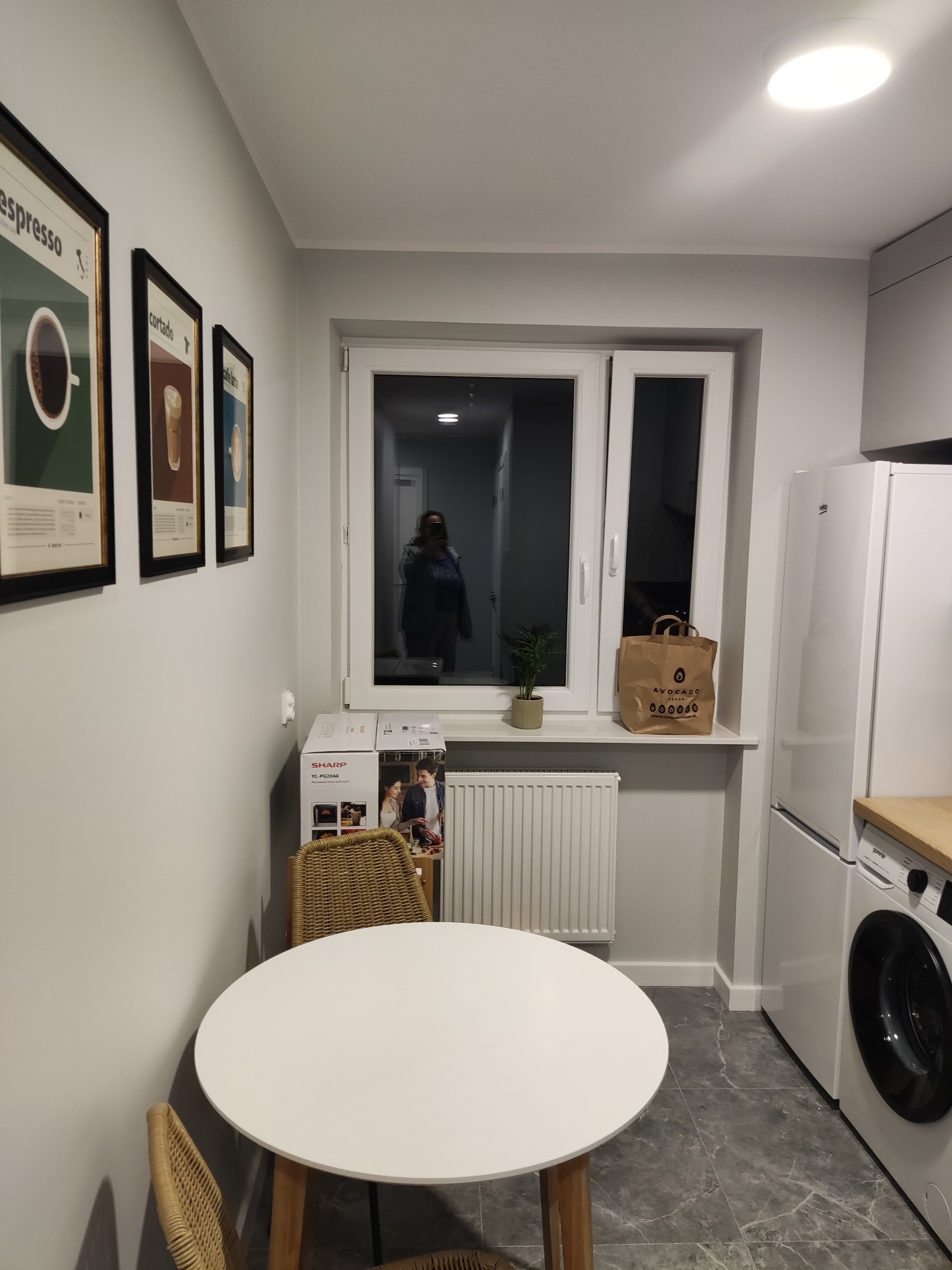 Wnętrze kuchni po remoncie: szare ściany, marmurowe płytki podłogowe, okrągły biały stół z wiklinowymi krzesłami, plakat na ścianie, torba na parapecie, widoczny nowy sprzęt AGD.