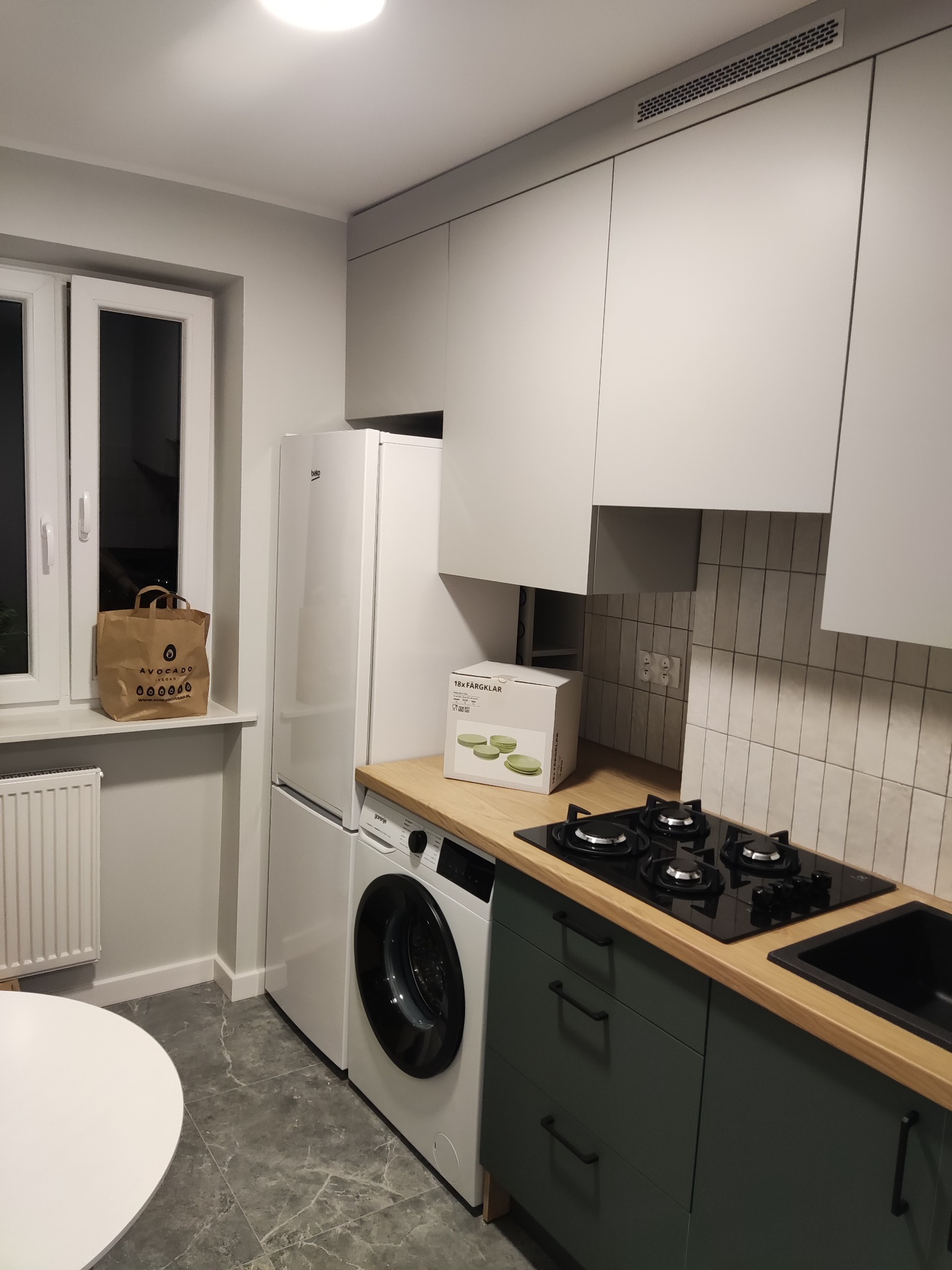 Nowoczesna kuchnia w odcieniach szarości i zieleni z drewnianym blatem, płytą grzewczą i zlewem, z pralką i lodówką w zabudowie, torba na oknie i pudełko z naczyniami na blacie.