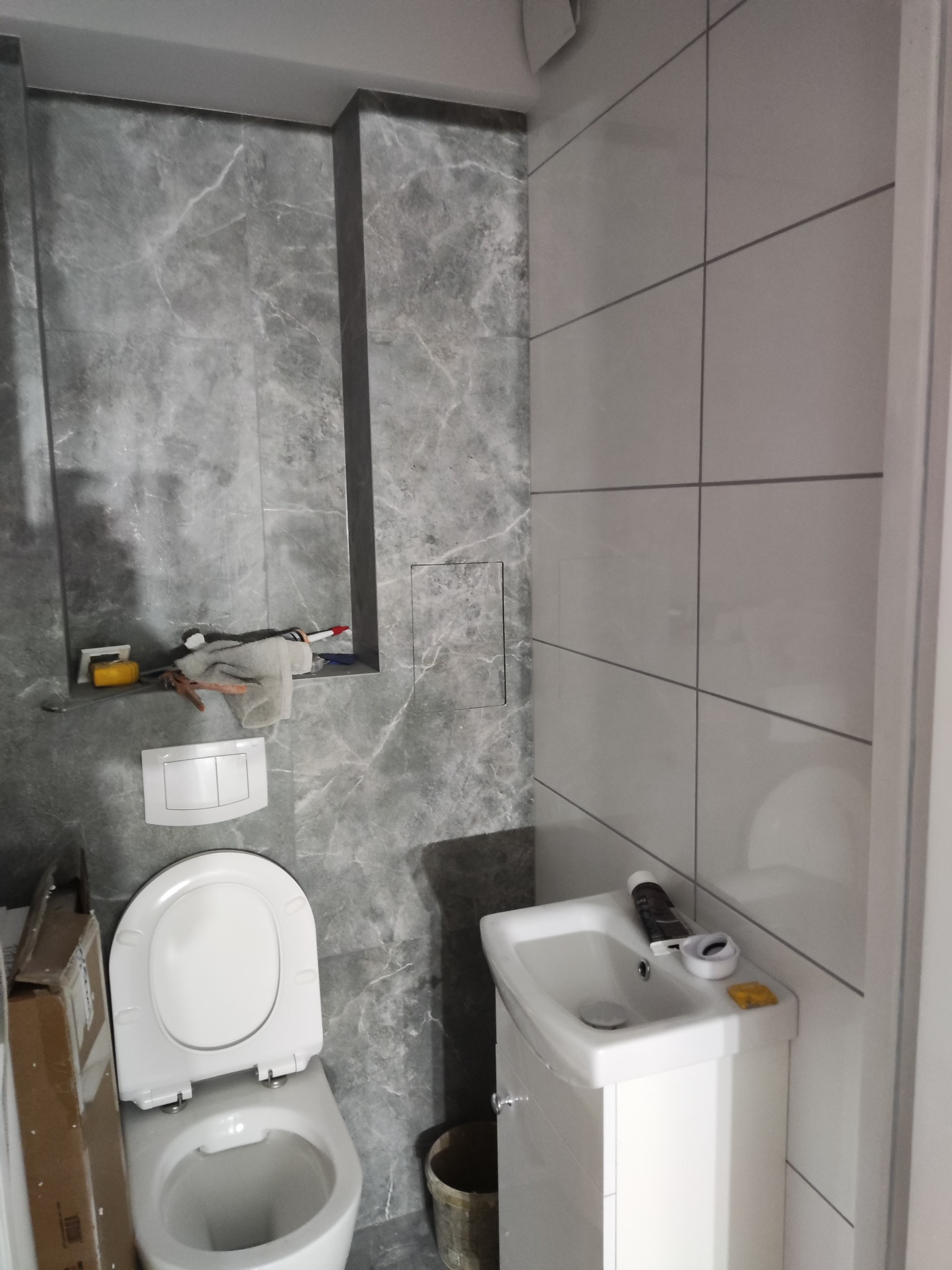 Łazienka w trakcie wykańczania: biała umywalka z szafką, toaleta oraz szare płytki imitujące marmur na ścianach, widoczne narzędzia i materiały budowlane.